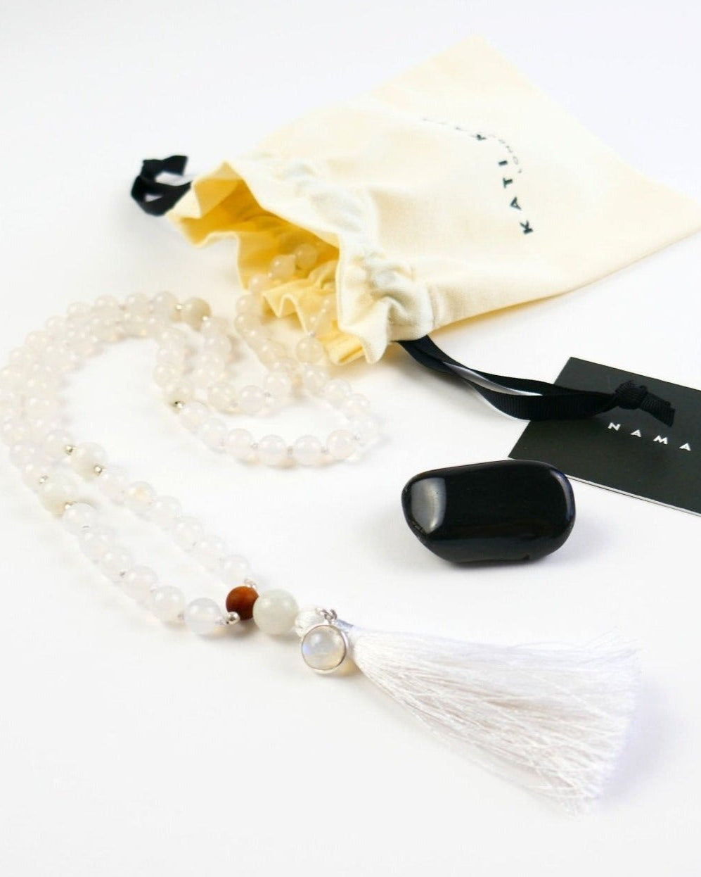 Kati Kaia crystal moonstone mala necklace with white beads and tassel next to cream pouch and black stone