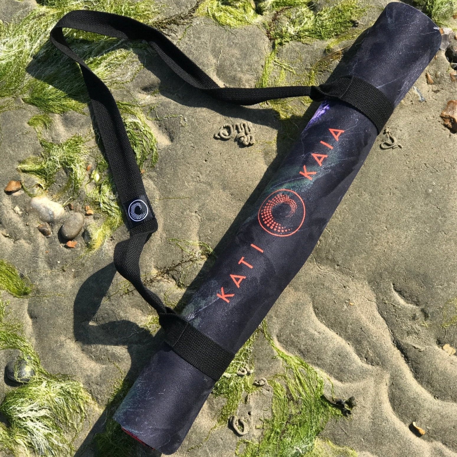 Kati Kaia portable yoga mat for travel rolled up with black carrying strap on sandy mossy beach
