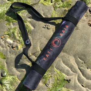 Kati Kaia portable yoga mat for travel rolled up with black carrying strap on sandy mossy beach