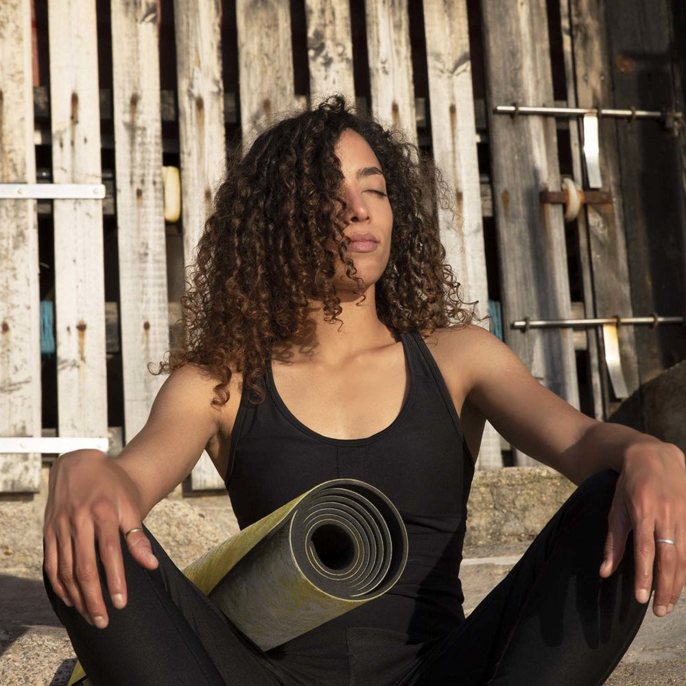 Woman sitting outdoors holding a rolled up yellow artist yoga mat by Kati Kaia in black workout wear